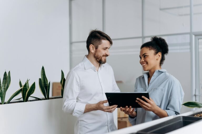 A black woman and a caucasian man discussing work with a clipboard in a modern office setting.